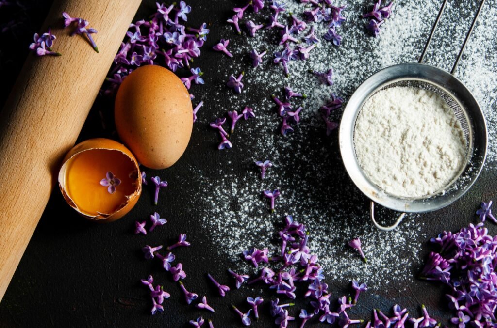 A rustic baking arrangement featuring eggs, flour, and lilac flowers on a dark surface with a sieve.