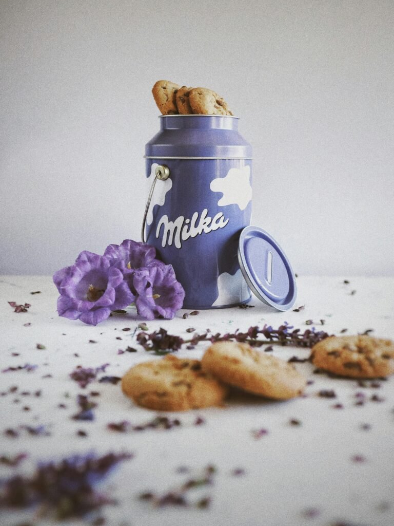 Delicious homemade cookies in a Milka tin surrounded by purple blooms on a white surface.
